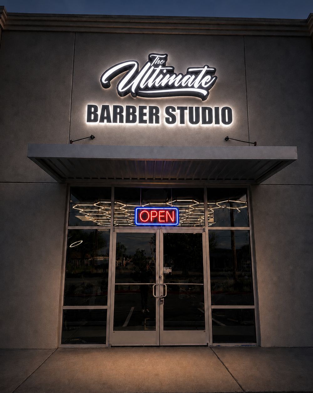 The Ultimate Barber Studio storefront at night: lit script sign, open sign, and hexagon ceiling lights through the glass